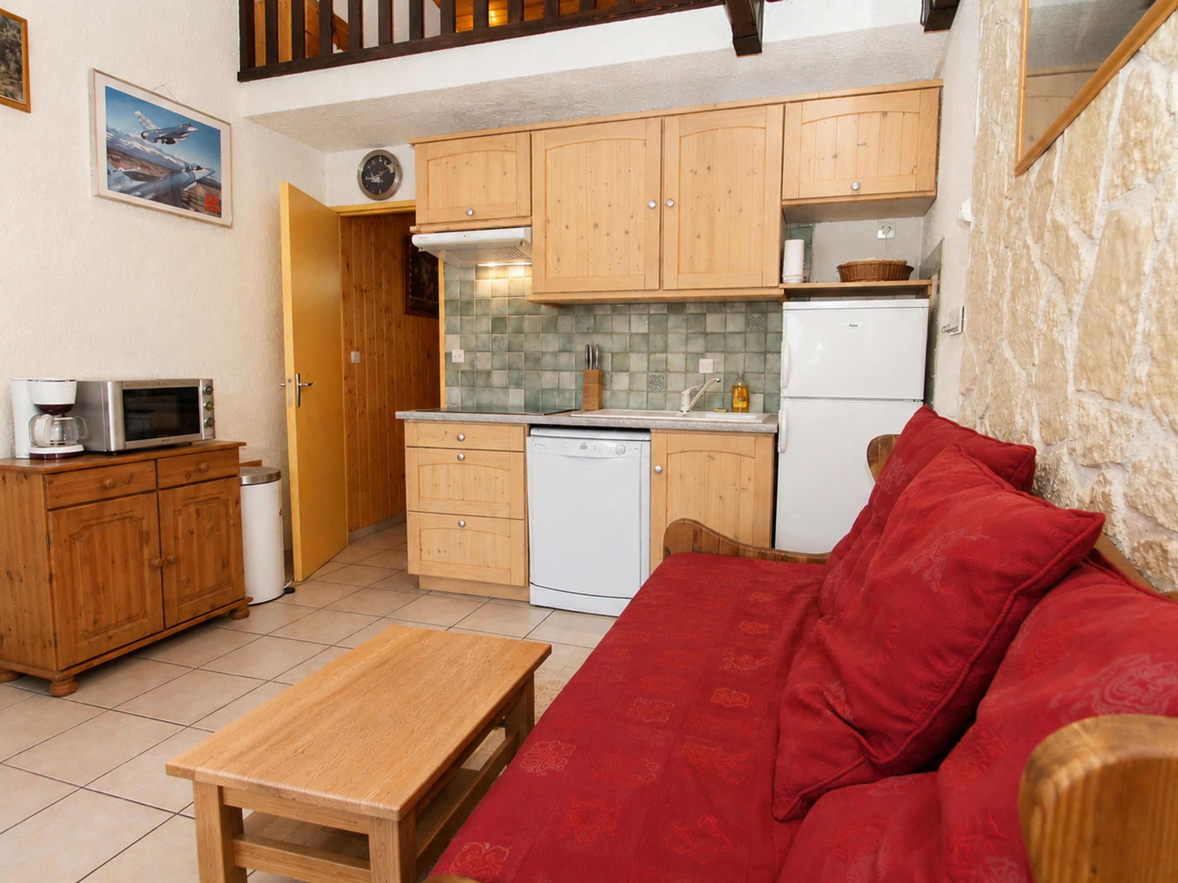 Een knus woongedeelte met een kitchenette, houten kastjes, een rode bank en een kleine houten tafel.