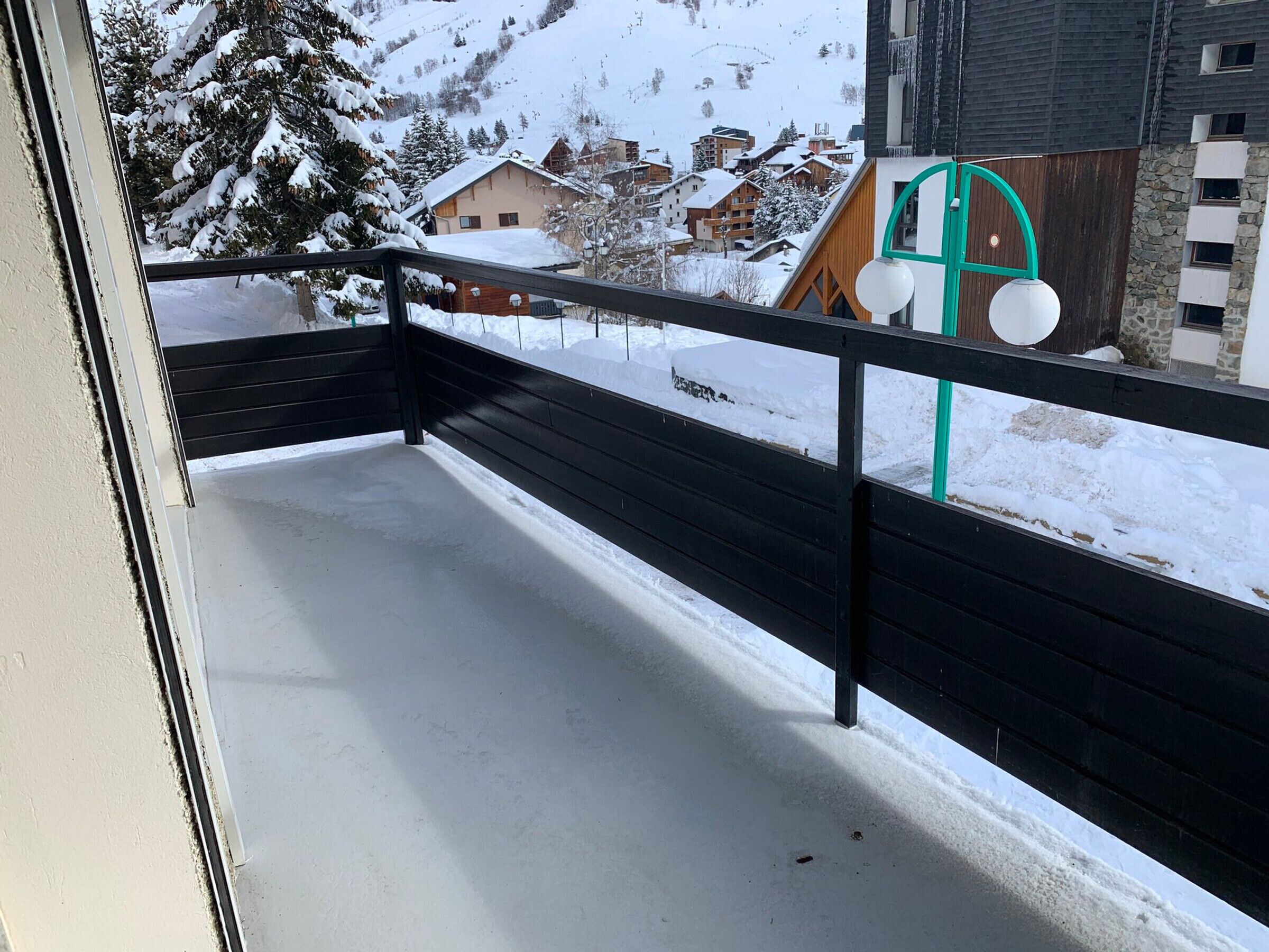 Een balkon met een met sneeuw bedekte grond, een houten hekwerk en een uitzicht op besneeuwde huizen en bomen op de achtergrond.