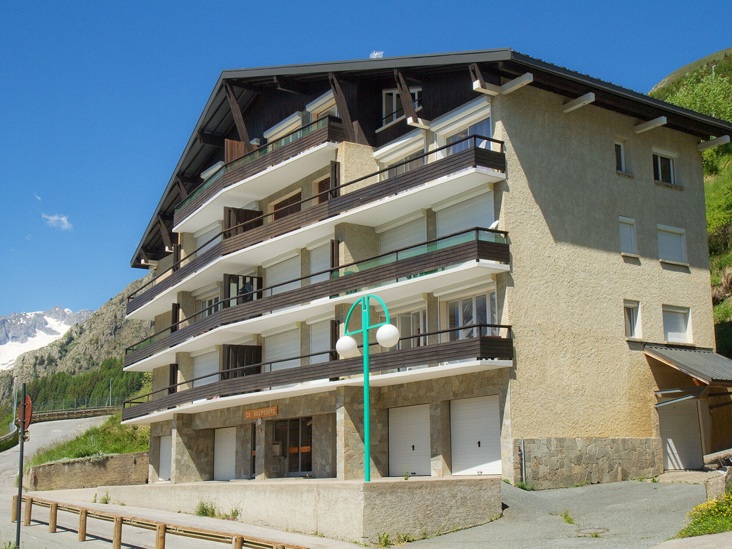 Un edificio de varios pisos con balcones, fachada de piedra y un paisaje montañoso de fondo.