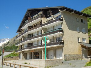Un immeuble à plusieurs étages, doté de balcons et d’une façade en pierre, avec une chaîne de montagnes en arrière-plan.