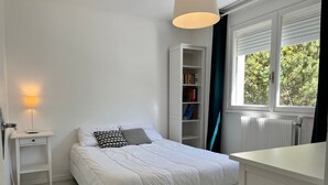 Un dormitorio con cama, mesita de noche, lámpara, estantería, ventana y radiador.