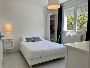 Ein Schlafzimmer mit Bett, Nachttisch, Lampe, Bücherregal, Fenster und Heizkörper.