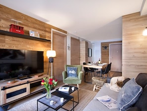 A modern living room with a flat-screen TV, a coffee table with a vase of flowers, a sofa with a cushion, and a dining area with a table set for two. - T2 Apartment, central and quiet - 4 guests (Les Deux Alpes)