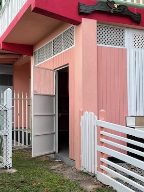 Un edificio con fachada rosa, cerca blanca y una puerta entreabierta que da a un espacio interior.
