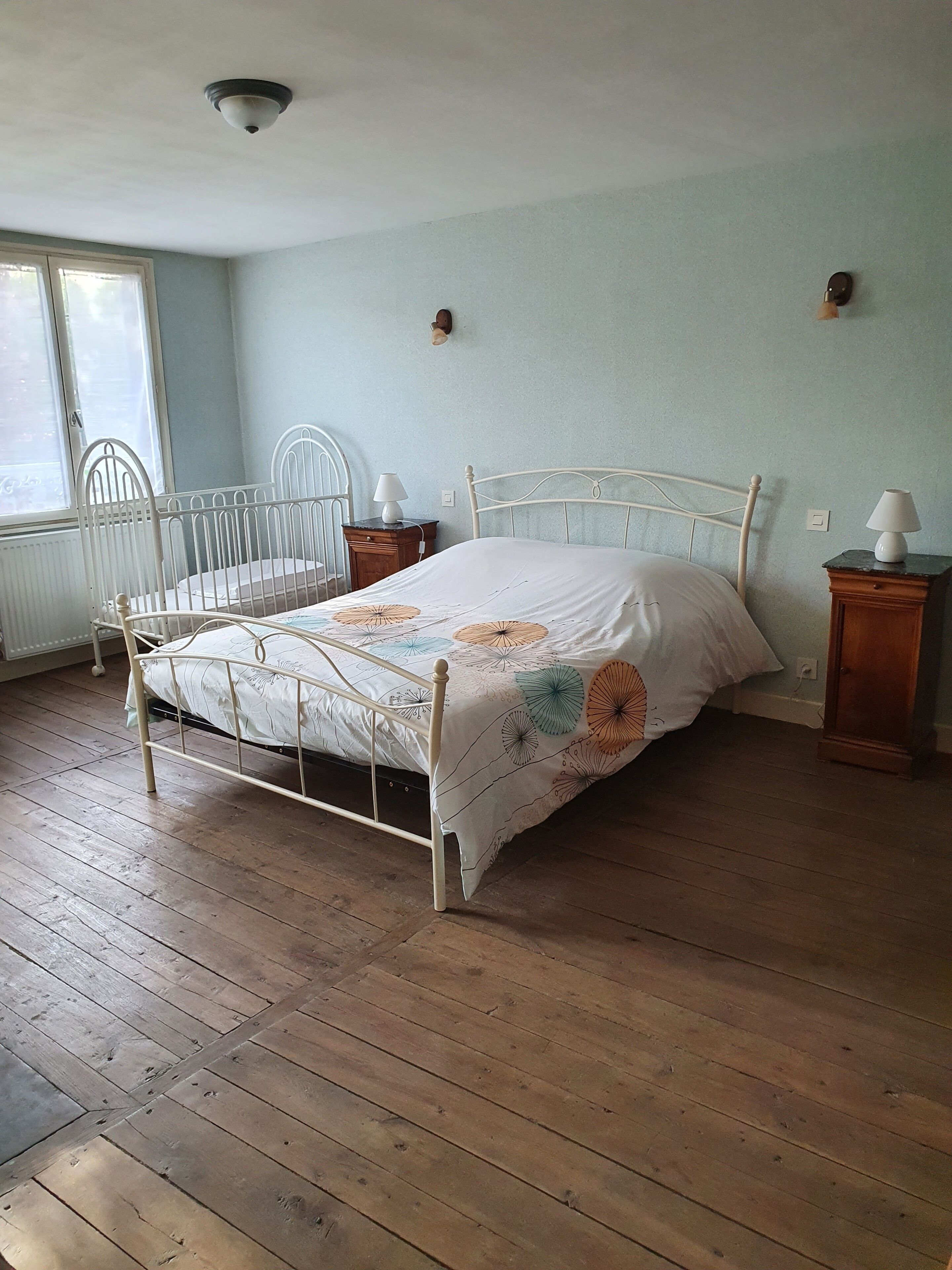 A bedroom with a metal-framed bed, a wooden floor, a window with blinds, and a wall-mounted light fixture.