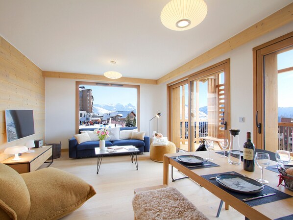 Un salon moderne avec un canapé, une table basse et une table à manger, le tout agrémenté de verres à vin et d’une bouteille. De grandes fenêtres offrent une vue sur un paysage montagneux enneigé.