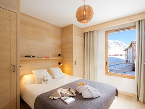 A bedroom with a wooden wall, a bed with pillows, a bedside table, a hanging light fixture, and a window with curtains. - Apartment, ski-in/ski-out, piste view (Huez)