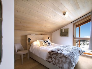 Ein Schlafzimmer mit Holzbalkendecke, einem Bett mit weißer Bettwäsche, einem Stuhl und einem Fenster mit Blick auf eine verschneite Landschaft.