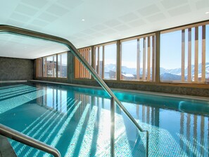An indoor swimming pool with clear blue water, a metal ladder, and large windows offering a view of snow-capped mountains. - Top floor apartment with mountain view - 4 guests (Huez)