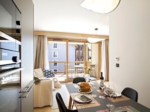 A modern living room with a dining area, a sofa, a table set for two, and a view of the outside balcony. - Bright apartment, ski-in/ski-out - 4 guests (Huez)
