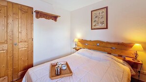 Une chambre à coucher avec une tête de lit en bois, une table de chevet avec une lampe, un lit recouvert d’une couette blanche, une armoire en bois et un plateau avec une bouilloire et des tasses.