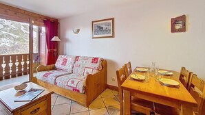 Une pièce chaleureuse avec un lit en bois, une table à manger dressée pour deux personnes et une fenêtre donnant sur un paysage enneigé.