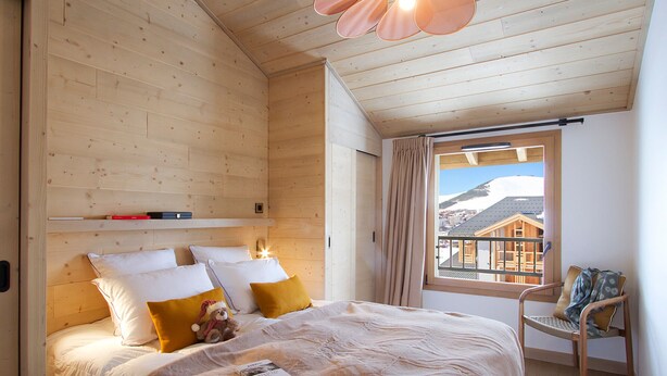 A wooden cabin bedroom with a bed, pillows, a teddy bear, a chair, and a window with a view of a mountain. - Spacious apartment, slope view - 10 guests (Huez)