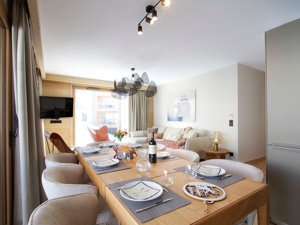 A modern dining area with a long wooden table set for four, a television mounted on the wall, and a sofa in the background. - Luxury apartment, station view - 6 guests (Huez)