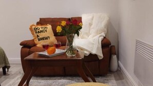 A living room with a brown sofa, a coffee table with a vase of flowers, and a yellow ottoman.