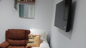 A living room with a brown armchair, a white sofa, a flat-screen TV mounted on the wall, and a small wooden coffee table.