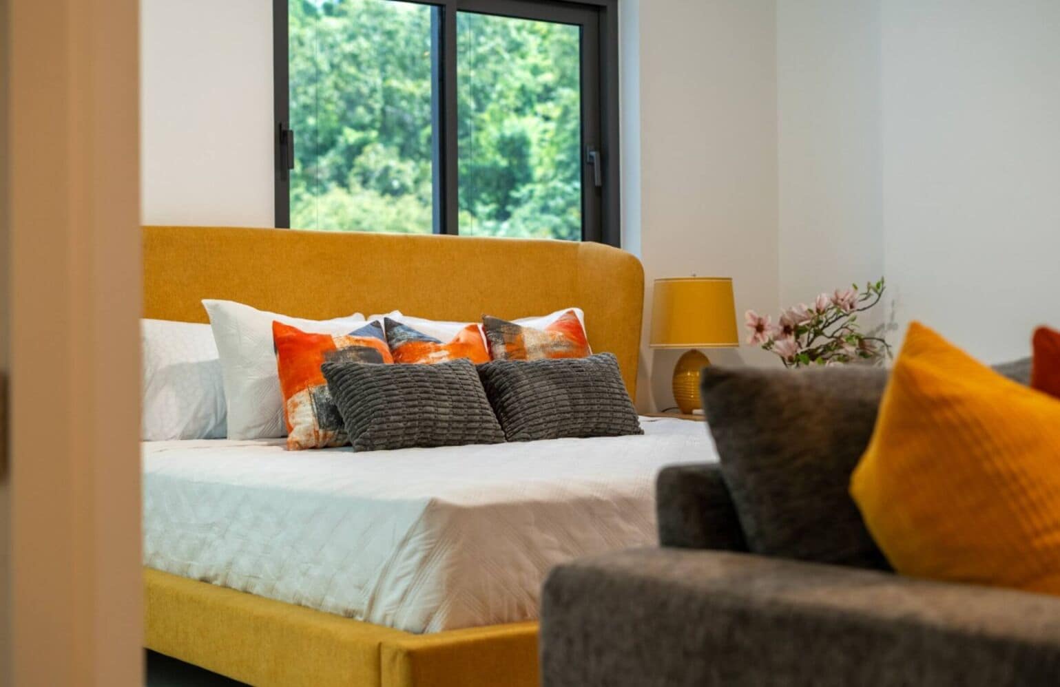 Una cama con varias almohadas, una mesita de noche con lámpara y una ventana con vistas al verdor.