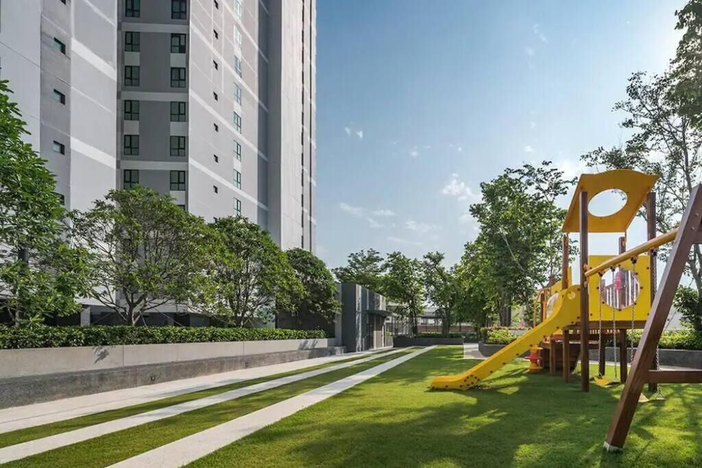 Un terrain de jeux avec un toboggan et des balançoires, entouré d’une pelouse bien entretenue et d’arbres, avec un grand immeuble en arrière-plan.