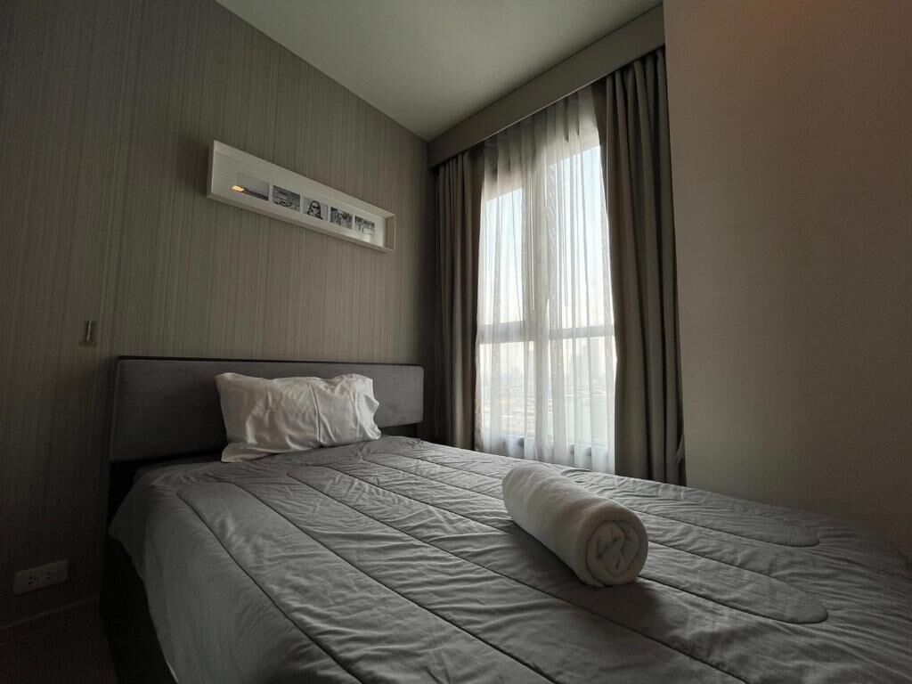 A hotel room with a bed, a headboard, a wall-mounted control panel, and a window with curtains.