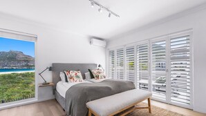 A bedroom with a large window, a bed, a bench, and a view of a coastal landscape. - PINGU Beachhouse | Beachfront Villa with Hot Tub, Ocean Views & Beach Access (Cape Town)