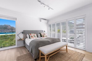 A bedroom with a large window, a bed, a bench, and a view of a coastal landscape. - PINGU Beachhouse | Beachfront Villa with Hot Tub, Ocean Views & Beach Access (Cape Town)