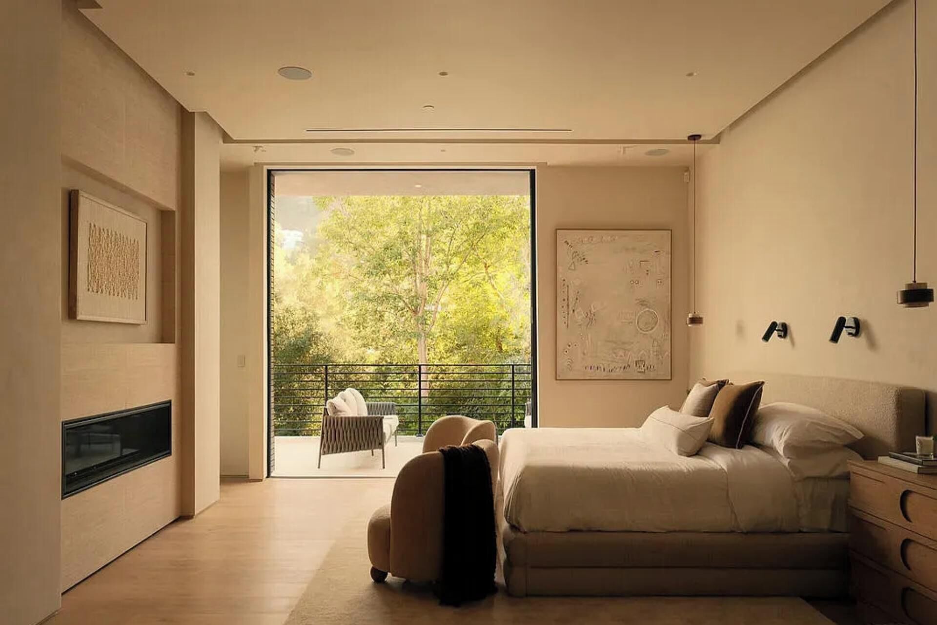 A modern bedroom with a bed, a chair, a fireplace, and a balcony with a view of trees.