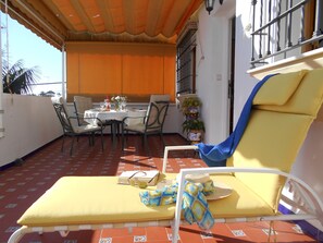 Eine sonnendurchflutete Terrasse mit einem Tisch für zwei Personen, einem gelben Liegestuhl und einem Blumentopf.