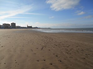 Ein weiter Sandstrand mit Fußspuren, die zum Wasser führen, ein entferntes Gebäude mit einem Turm und ein klarer Himmel.