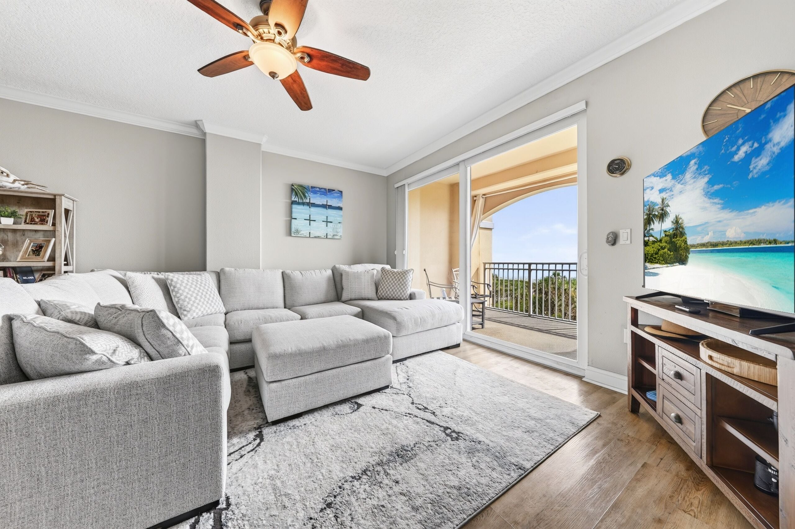 A living room with a sectional sofa, a ceiling fan, a flat-screen TV, and a view of a balcony with a railing.