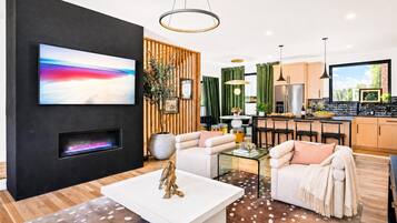 A modern living room with a flat-screen TV, a fireplace, a sofa, a coffee table, and a kitchen area with a refrigerator and bar stools.