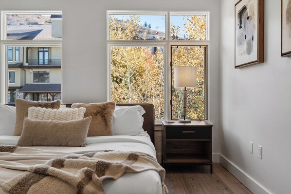 A bedroom with a bed, pillows, a bedside table with a lamp, and a window with a view of trees and a building. - Luxury Penthouse Studio | Ski-In/Ski-Out | Canyons Village Park City (Park City)