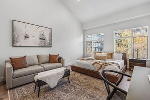 Een moderne slaapkamer met een groot bed, een bank, een bureau en een stoel. Er is een raam met uitzicht op bomen en een schilderij aan de muur.