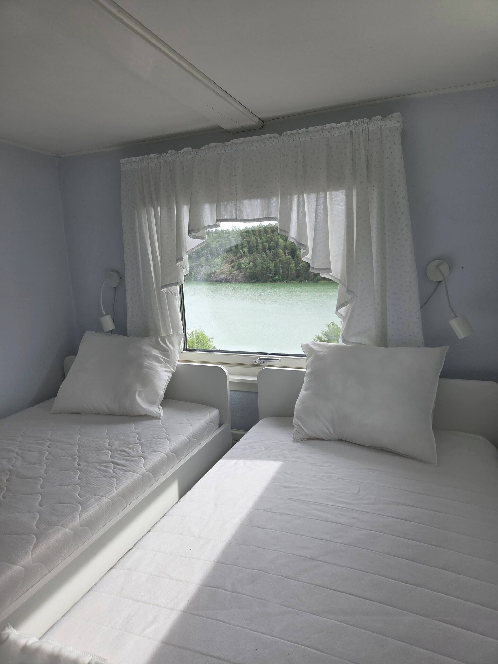 Ein Zimmer mit zwei Betten, einem fensterbelaubten Fenster und Blick auf ein Gewässer und Bäume.