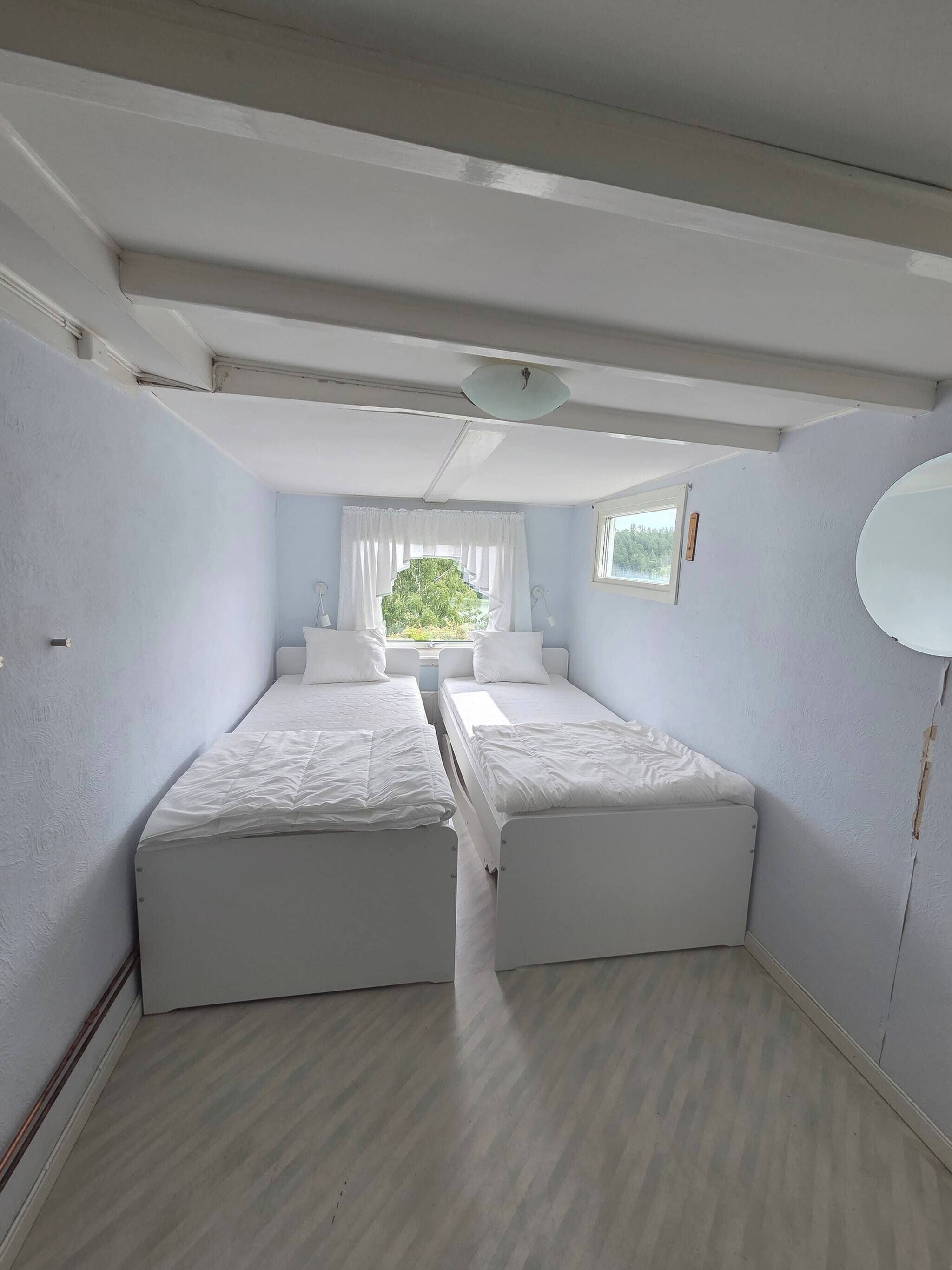 A room with two beds, a window with curtains, a ceiling light, and a view of greenery outside.