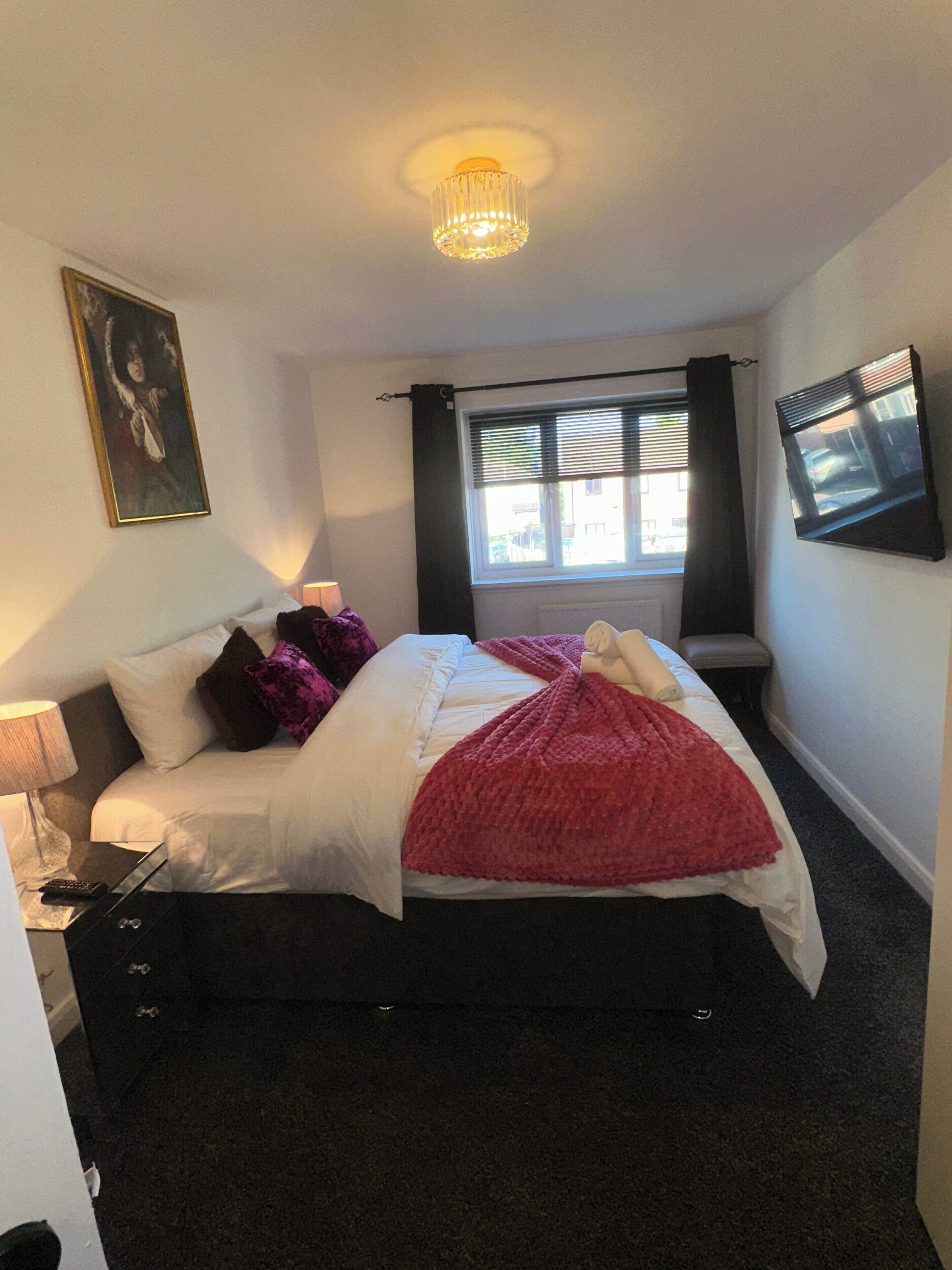 A bedroom with a bed, a bedside table, a lamp, a window with curtains, a painting on the wall, and a ceiling light fixture.