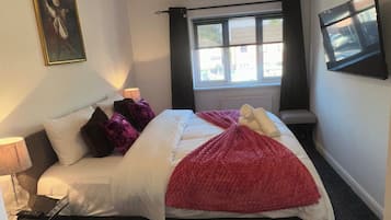 A bedroom with a bed, a bedside table, a lamp, a window with curtains, a painting on the wall, and a ceiling light fixture.