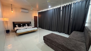 Una habitación de hotel con cama, sofá, lámpara de pie, aire acondicionado y ventana con cortinas.