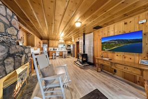 A cozy cabin interior with a stone fireplace, wooden walls, a large TV, and a kitchen area. - 3BR Tahoe Hideaway with Mountain Views and Rustic Feels (South Lake Tahoe)