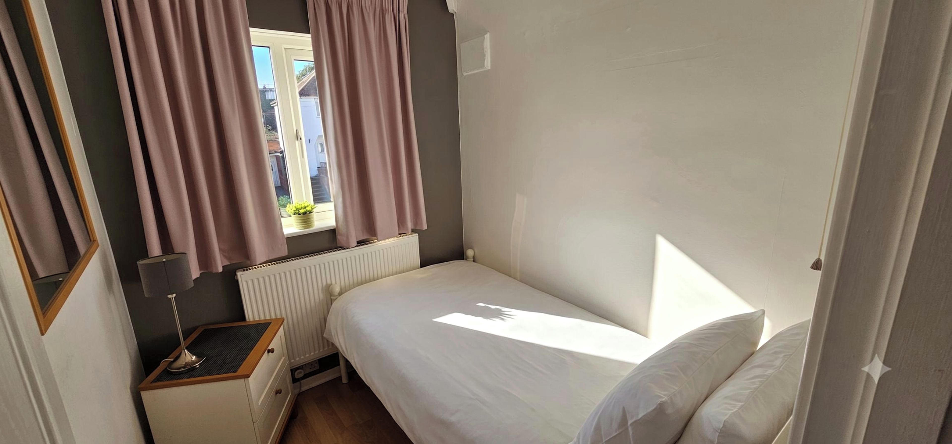 A small, single-bed room with a window, a radiator, a bedside table, and a lamp.