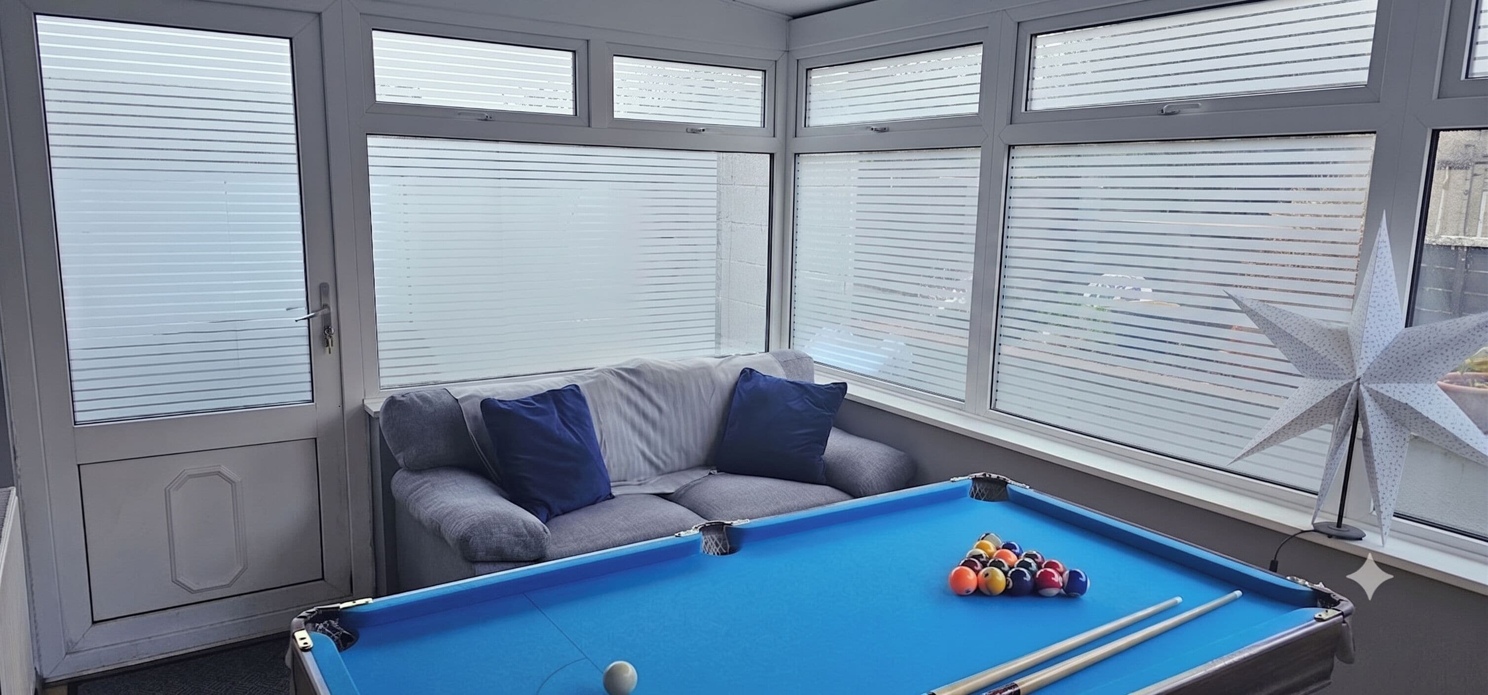 Ein Billardtisch mit Kugeln und Spielstöcken, ein graues Sofa mit blauen Kissen und ein Fenster mit Jalousien.