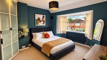 A bedroom with a bed, a window with curtains, a radiator, and a view of houses outside.