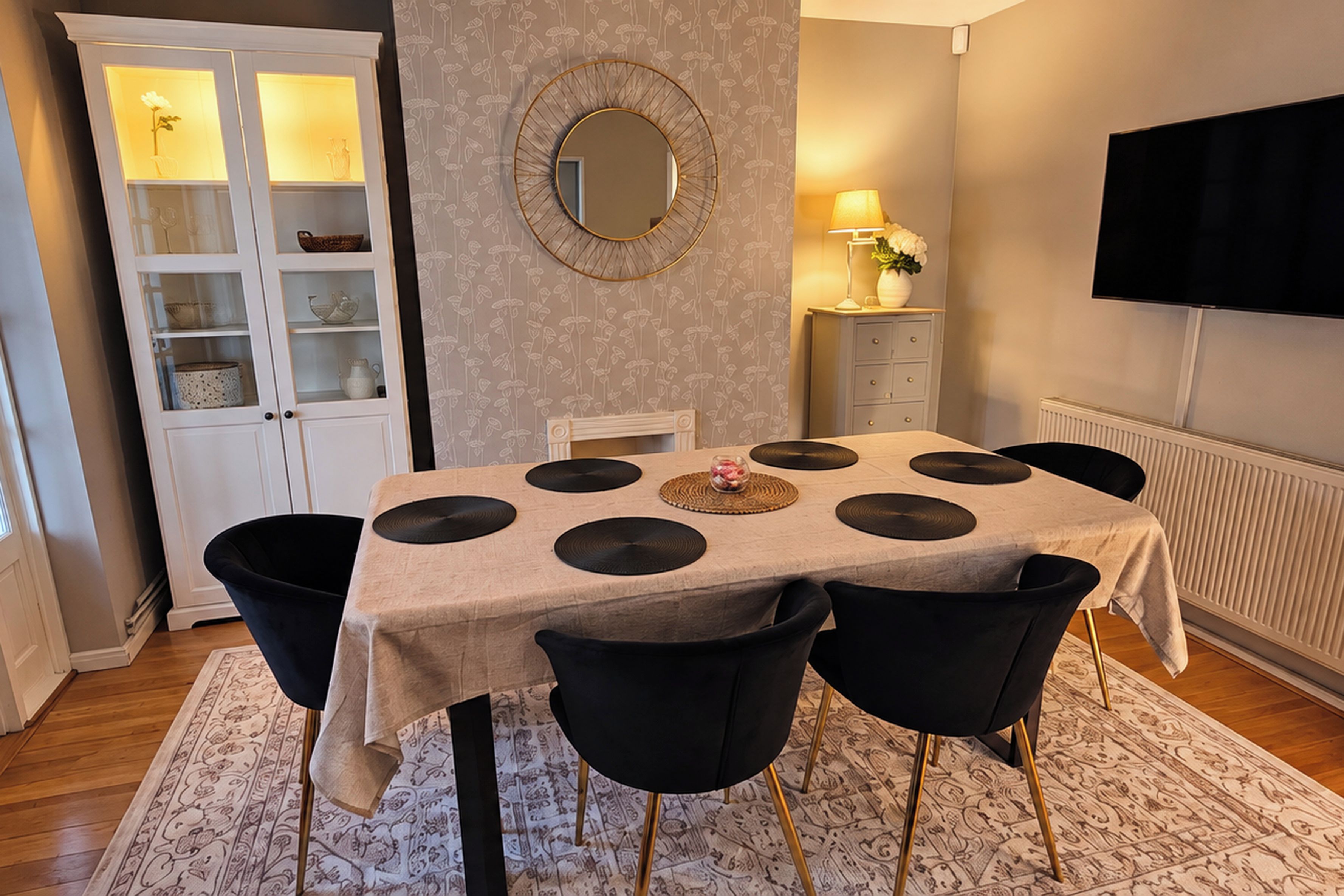 A dining area with a table, chairs, a cabinet, a mirror, a lamp, and a television on the wall.