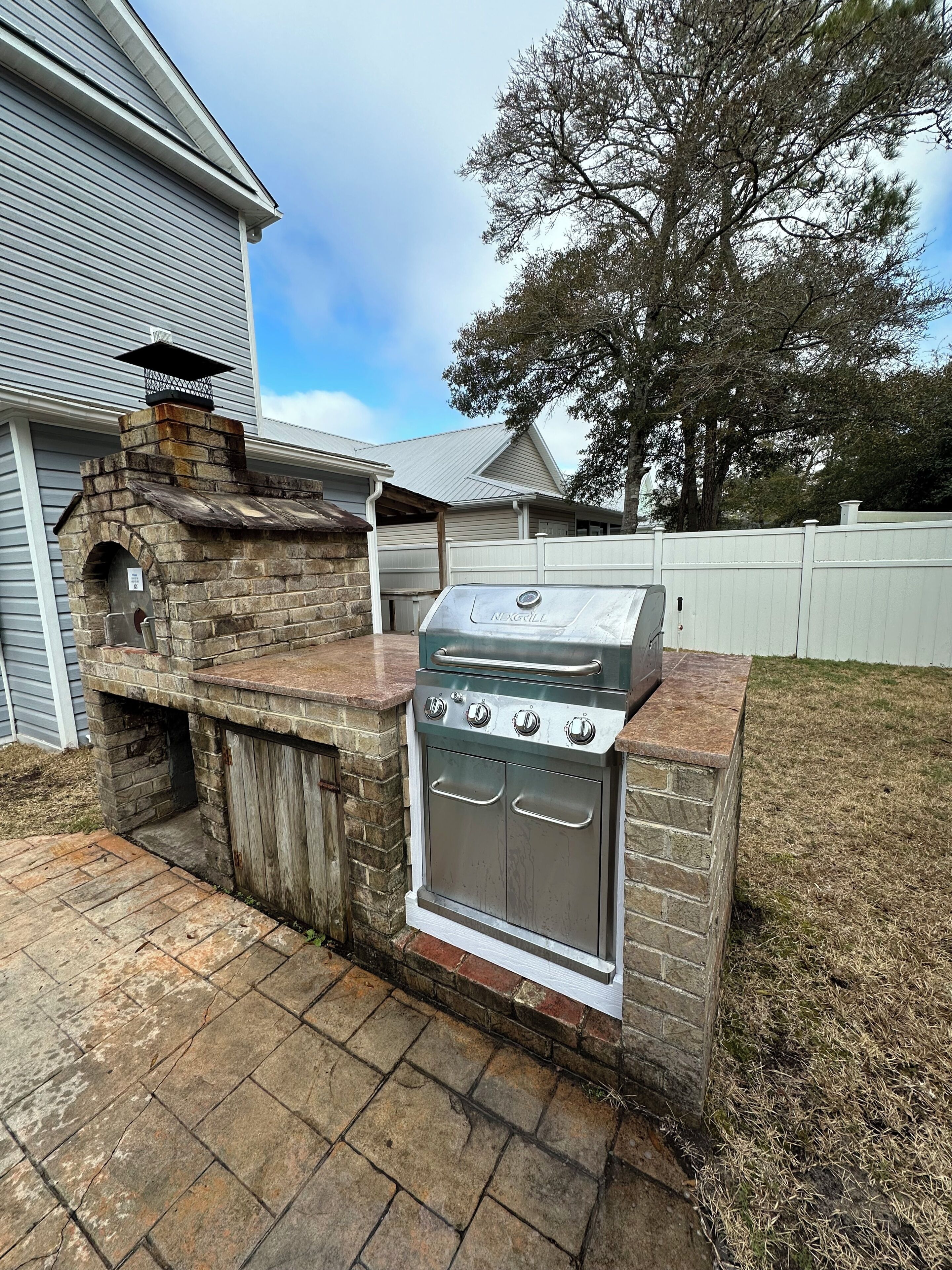 A stone-built outdoor kitchen with a grill, adjacent to a house with a white fence.