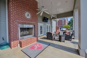 A covered patio with a brick fireplace, outdoor seating, ceiling fans, and a TV mounted on the wall. - Tusk & Tide | Near the Stadium & Campus! (Tuscaloosa)