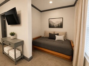 Ein Schlafzimmer mit einem Bett, einem an der Wand montierten Flachbildfernseher, einem Nachttisch mit einer Pflanze und einem Fenster mit Vorhängen.