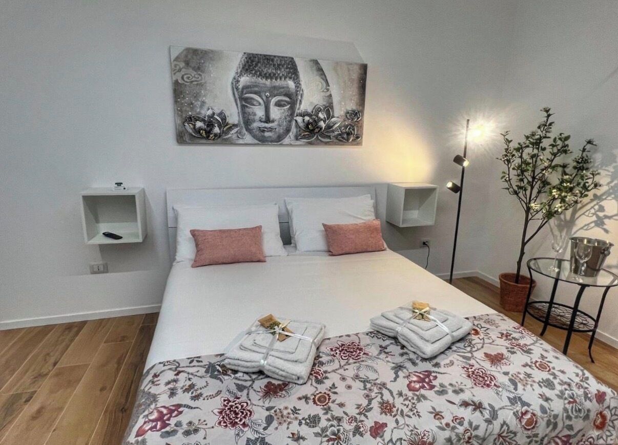A hotel room with a bed, pillows, a nightstand, a wall-mounted artwork, a floor lamp, and a potted plant.