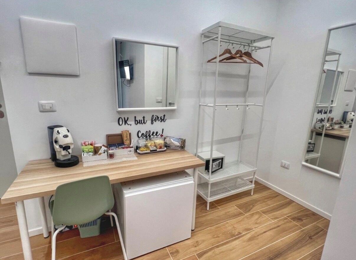A compact room with a desk, a chair, a small refrigerator, a mirror, and a shelving unit with hangers.