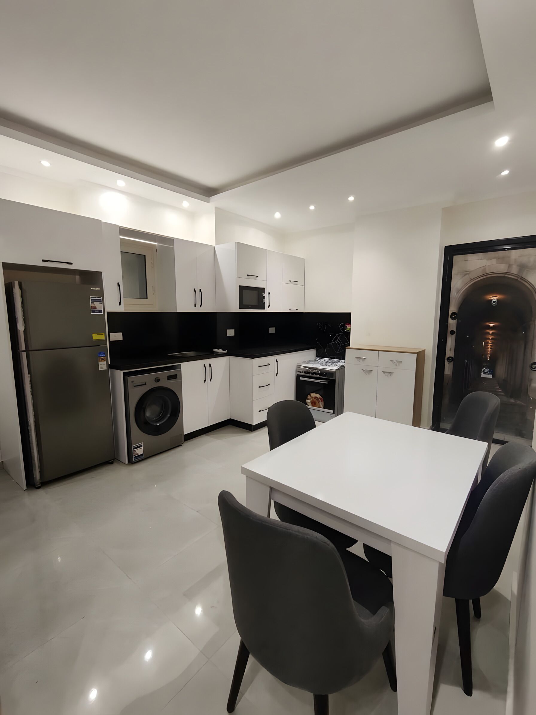 Eine moderne Küche mit weißen Schränken, einer schwarzen Arbeitsplatte, einem Edelstahlkühlschrank, einer Waschmaschine und einem Essbereich mit weißem Tisch und schwarzen Stühlen.