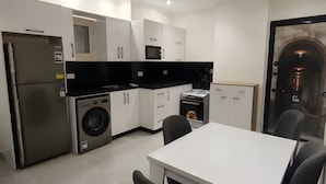 Eine moderne Küche mit weißen Schränken, einer schwarzen Arbeitsplatte, einem Edelstahlkühlschrank, einer Waschmaschine und einem Essbereich mit weißem Tisch und schwarzen Stühlen.
