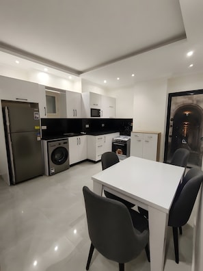 A modern kitchen with white cabinets, a black countertop, a stainless steel refrigerator, a washing machine, and a dining area with a white table and black chairs. - Hurghada Hub 2BR Pool View Apartment with Aqua Park Access (Hurghada)